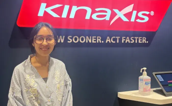 Photo of Kinaxis intern Yui Nagasaka standing next to a wall with the red Kinaxis logo sign