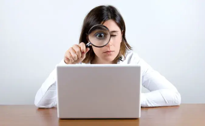 Woman in white shirt peers through a magnifying glass at a laptop computer