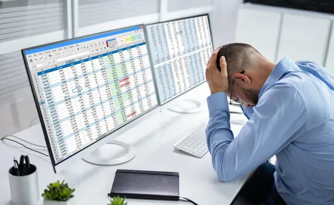 Man sits at a desk in front of two computer screens displaying spreadsheets. He is holding his head in a manner that indicates stress or defeat.