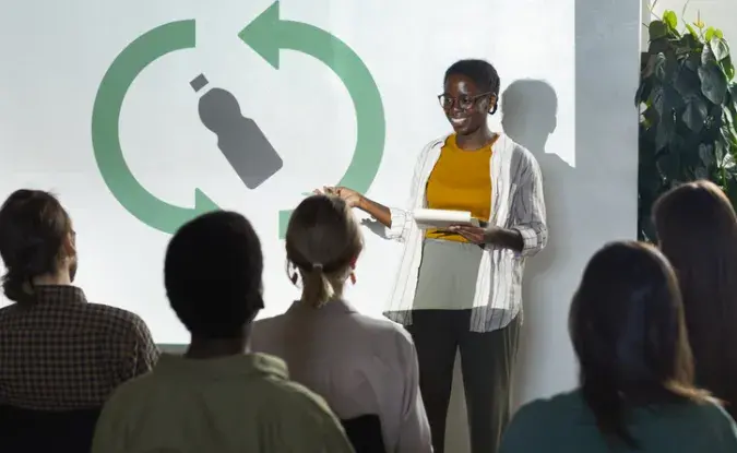 Business woman presenting a slide to a small audience. On the slide is a graphic of a bottle with a recycle logo surrounding it.