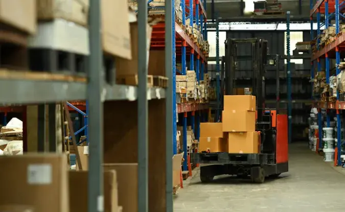A forklift holding a messy stack of boxes is parked in a warehouse lined with full shelves.