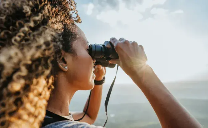 A woman looks out of binoculars towards a bright horizon.