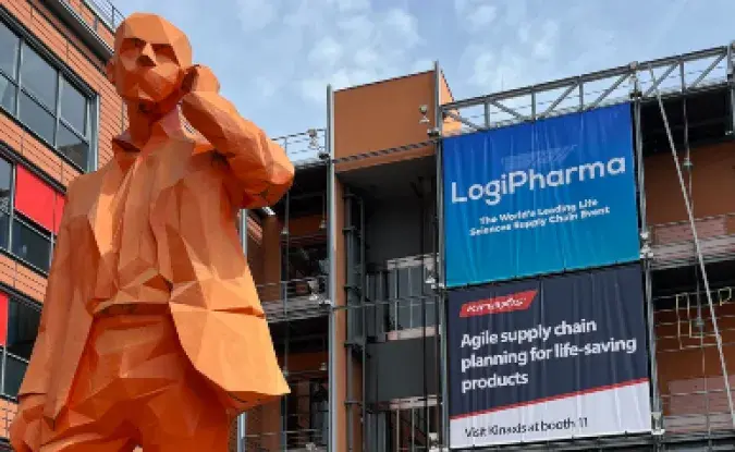 A large orange-colored sculpture of a man outside a conference center in Lyons, France with building signage reading LogiPharma 2023 with the red Kinaxis logo.