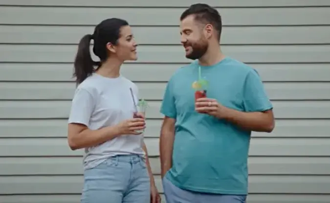 A woman in a white t-shirt and jean shorts stands next to man in green t-shirt and jeans, holding summer cocktails and smiling at one another.