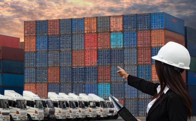 Female engineer wearing hard hat working with container cargo freight ship and trucks in shipyard at dusk.