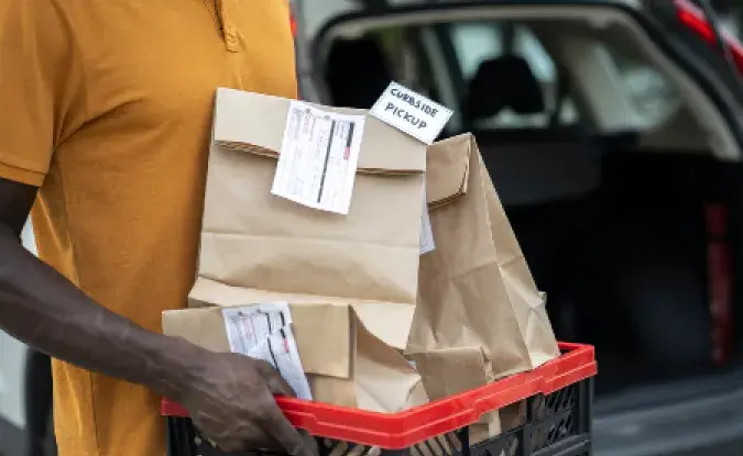 Close up of a curbside pickup food delivery. Service staff holding a basket with food delivery, standing behind a car trunk.