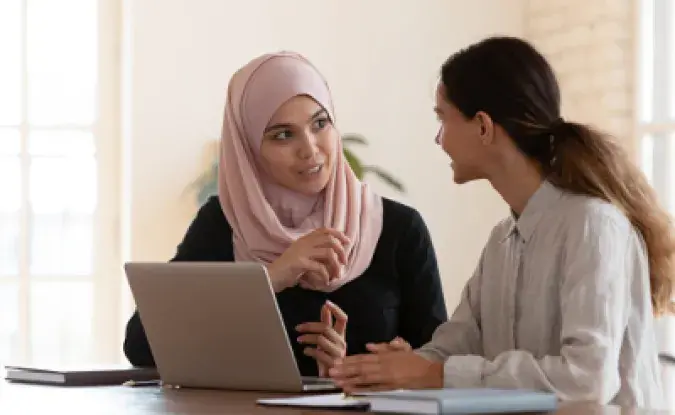 A woman in a pale pink hijab sits in front of an open laptop computer. A woman with long brown hair in a ponytail on the right sits next to her and they are talking.