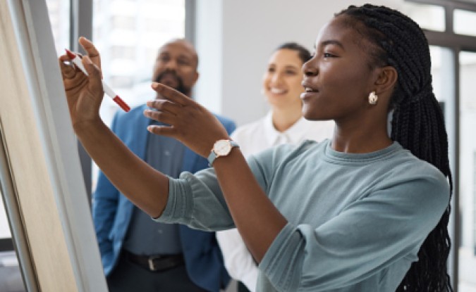 Young black woman writes on a whiteboard at work while two people of different races are in background, smiling toward her.