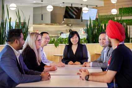 A group of diverse Kinaxis employees sit together at a table in the Kinaxis common space and cafeteria, smiling and having a discussion.