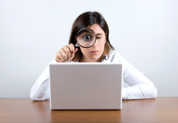 Woman in white shirt holds magnifying glass up to one eye as she stares at a laptop computer.