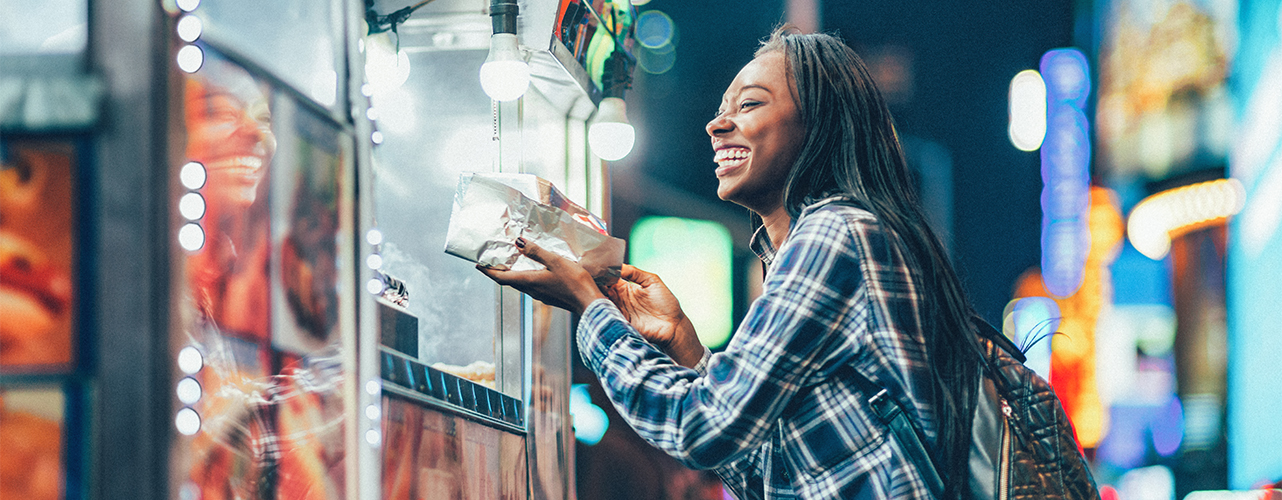 A woman receives a hot dog from a street vendor, representing a happy customer when the supply chain planning relationship for hot dog items remains intact.