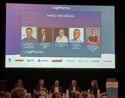 An audience viewpoint of the LogiPharma stage during a panel discussion with five individuals seated along the bottom of the image, and a large projected screen displaying a presentation slide behind and above them.