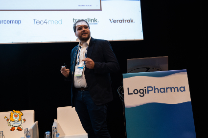 ​ EQRx exec Lou Lozanov onstage next to a podium reading "LogiPharma" in front of a projected slide show during his presentation at LogiPharma 2023