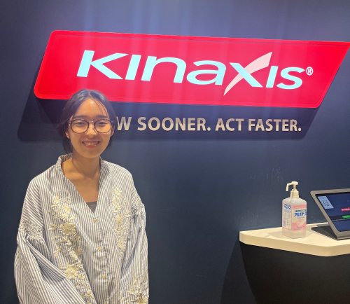 Photo of Kinaxis intern Yui Nagasaka standing next to a wall with the red Kinaxis logo sign