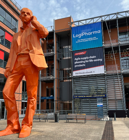 A large orange-colored sculpture of a man outside a conference center in Lyons, France with building signage reading LogiPharma 2023 with the red Kinaxis logo.